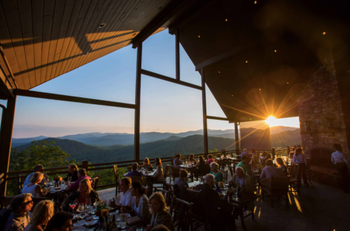 A restaurant with large windows overlooking mountains at sunset, filled with diners enjoying the view.