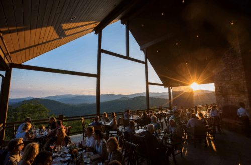 A restaurant with large windows overlooking mountains at sunset, filled with diners enjoying the view.