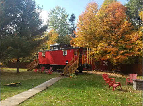 A red caboose converted into a cabin, surrounded by colorful autumn trees and a grassy area with Adirondack chairs.