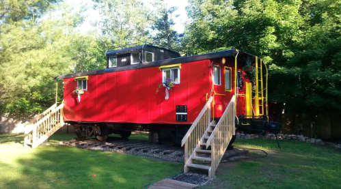 A bright red caboose on a grassy area, featuring wooden steps and surrounded by trees.