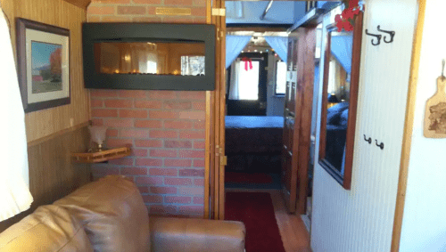 Cozy interior of a tiny home featuring a brick wall, a sofa, and a view into a bedroom area. Warm lighting adds charm.