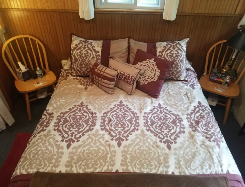 A neatly made bed with decorative pillows, surrounded by two wooden chairs and a lamp on a bedside table.