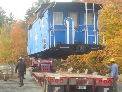 A blue caboose is being lifted onto a flatbed truck, surrounded by autumn trees and workers in hard hats.