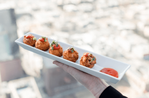 A hand holds a long white plate with five golden-brown croquettes topped with sauce and garnished, against a city backdrop.