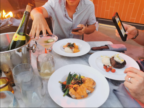 A table set with gourmet dishes, drinks, and a person taking a photo of their meal in a warm, outdoor setting.