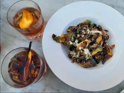 A plate of crispy Brussels sprouts topped with cheese, accompanied by two cocktails with orange garnishes.