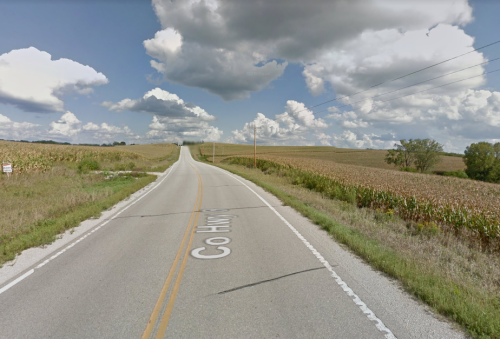 A rural road stretches into the distance, flanked by cornfields under a partly cloudy sky.