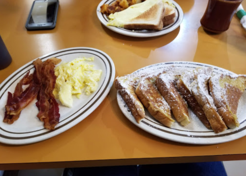 Two plates of breakfast: one with bacon and scrambled eggs, the other with French toast dusted with powdered sugar.