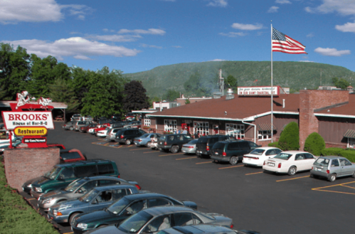 A restaurant with a parking lot, surrounded by trees and mountains, featuring an American flag and a sign for Brooks BBQ.
