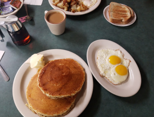 A plate with two pancakes topped with butter and syrup, alongside two sunny-side-up eggs and a cup of coffee.