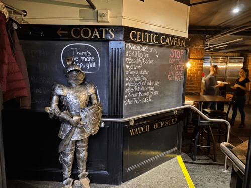 A knight statue stands near a chalkboard menu in a cozy pub setting with people in the background.
