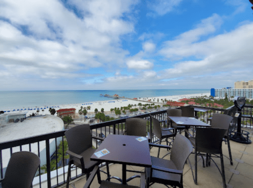 View from a balcony overlooking a beach with palm trees, blue water, and a cloudy sky. Tables are set for dining.