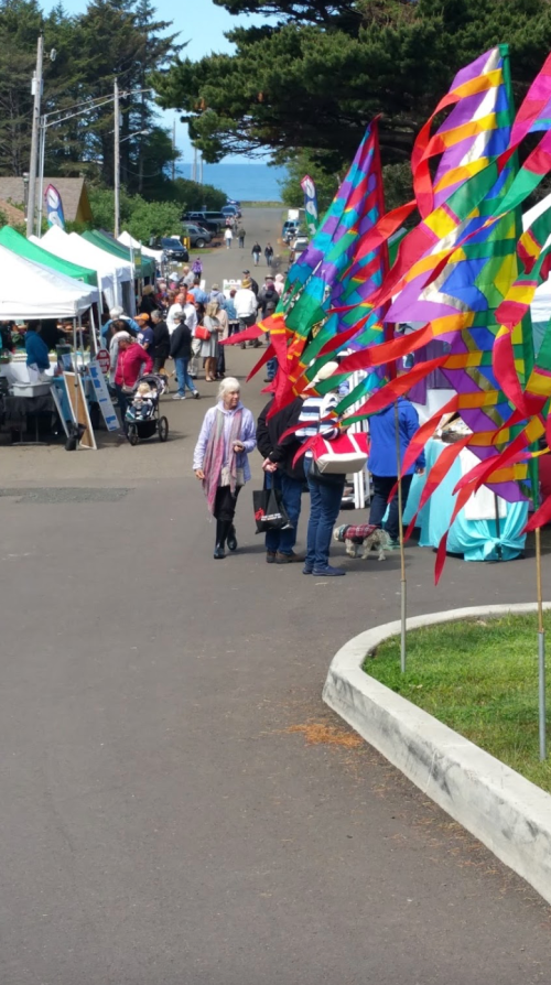 A vibrant street fair with colorful flags, tents, and people walking along a sunny path lined with trees.