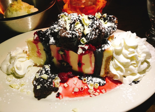 A decadent dessert featuring layers of cheesecake, chocolate sauce, whipped cream, and a cherry topping.
