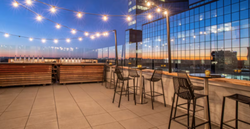 Rooftop bar with string lights, wooden counters, and high chairs, overlooking a city skyline at sunset.
