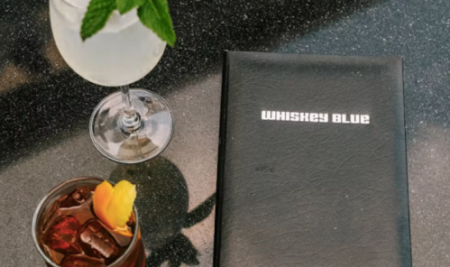 A cocktail and a whiskey drink beside a menu titled "Whiskey Blue" on a dark tabletop.
