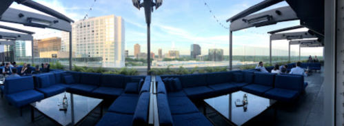 Panoramic view of a rooftop lounge with blue seating and city skyline in the background under a clear sky.