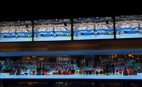 A bar with a well-stocked shelf of bottles, featuring multiple screens showing a tennis match.