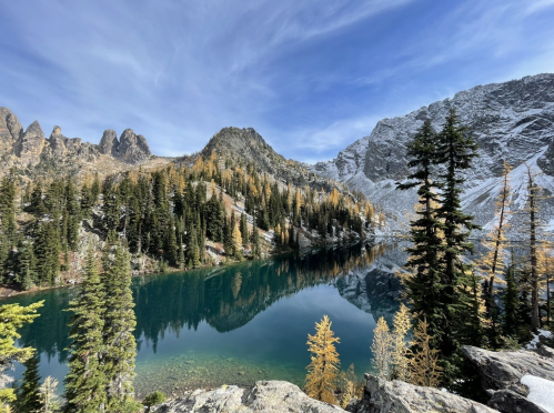 A serene mountain lake surrounded by evergreen trees and golden larches, with rocky peaks under a blue sky.