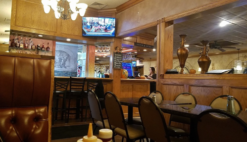 Interior of a restaurant featuring wooden decor, a bar area, and a TV, with empty tables and chairs in the foreground.