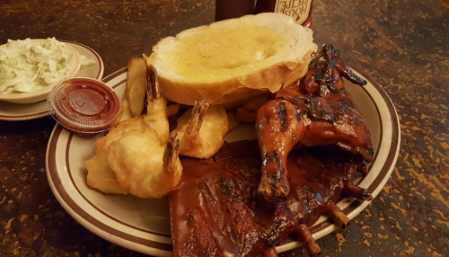 A plate featuring ribs, fried shrimp, a slice of buttered bread, and a side of coleslaw with dipping sauce.
