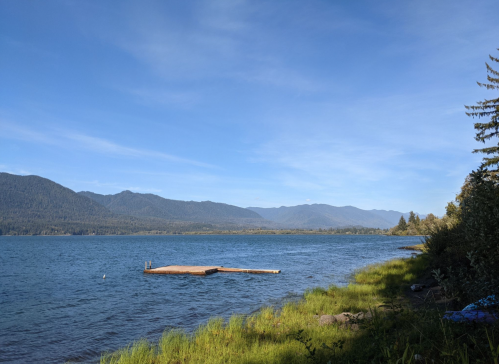 A serene lake view with a wooden dock, surrounded by green grass and mountains under a clear blue sky.