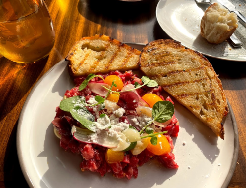 A plate of beef tartare topped with cherry tomatoes, herbs, and crumbled cheese, served with toasted bread.