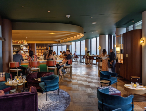 A stylish bar with patrons seated at tables and the counter, featuring modern decor and large windows with a city view.
