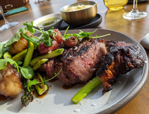 A plate of grilled steak with asparagus, roasted tomatoes, and microgreens, served with sides in the background.