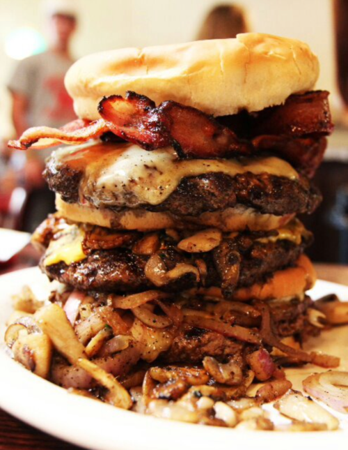 A towering burger with multiple beef patties, melted cheese, bacon, and sautéed mushrooms, served on a plate.