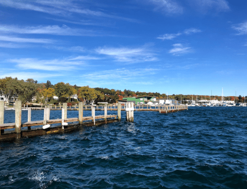 A wooden pier extends into a blue lake, with colorful trees and boats in the background under a clear sky.