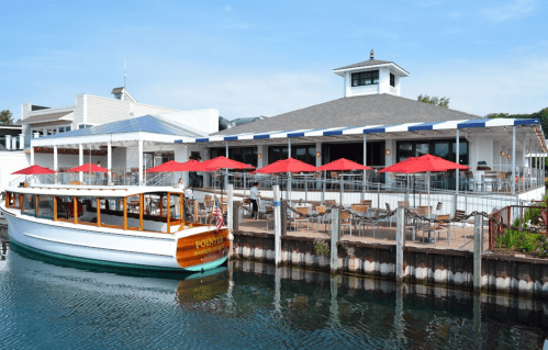 A waterfront restaurant with outdoor seating and red umbrellas, alongside a boat docked at the pier.