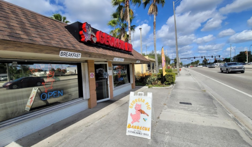 Exterior of Georgia Pig Barbecue restaurant with a sign and palm trees, located on a busy street under a blue sky.