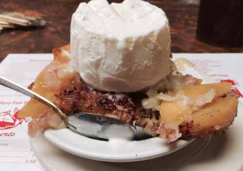A warm peach cobbler topped with a scoop of vanilla ice cream, served in a small dish with a spoon.
