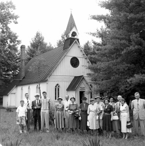 A group of people poses in front of a small church surrounded by trees, dressed in vintage clothing.