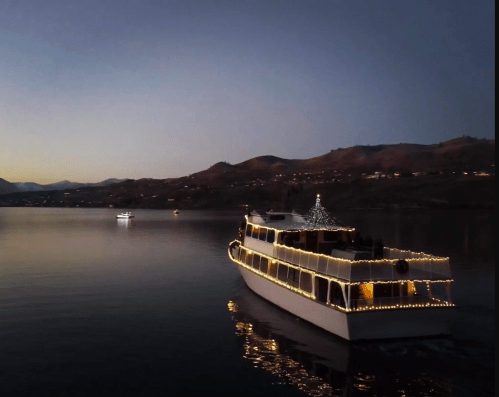 A beautifully lit boat glides across calm waters at dusk, with mountains in the background and another boat in the distance.