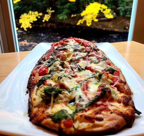 A flatbread pizza topped with spinach, tomatoes, and cheese, served on a white plate with a scenic background.