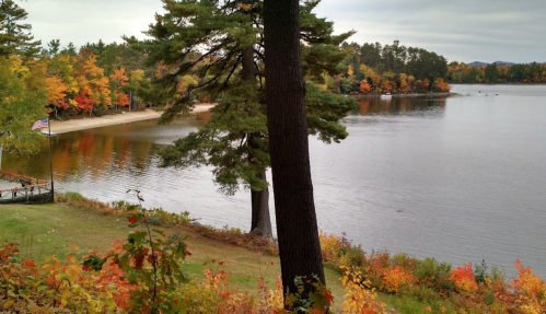 A serene lake surrounded by autumn foliage, with a sandy beach and a flag in the foreground.