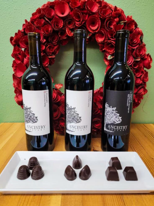 Three bottles of Ancestry wine in front of a red rose wreath, accompanied by a plate of assorted dark chocolates.