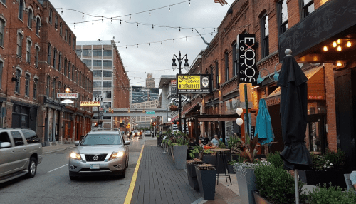 A lively street scene with restaurants, string lights, and a car driving by in an urban setting.