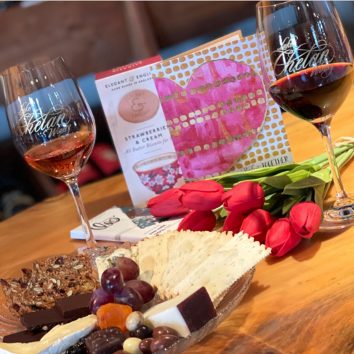 Two glasses of red wine beside a plate of assorted chocolates and cheeses, with tulips and a greeting card in the background.