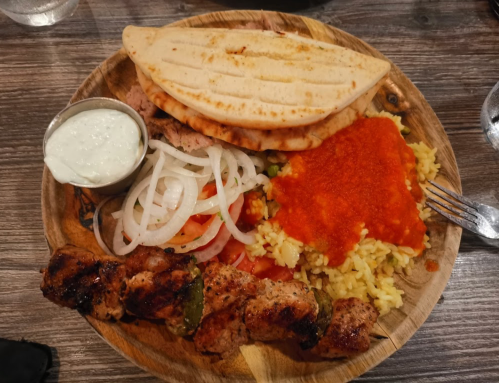 A wooden plate with grilled meat skewers, rice, salad, pita bread, and a small bowl of sauce.