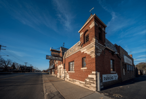 A brick building with a tower and sign reading "Rathseller," located at a street corner under a clear blue sky.