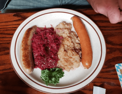 A plate with two sausages, red cabbage, mashed potatoes, and a piece of lettuce on a wooden table.