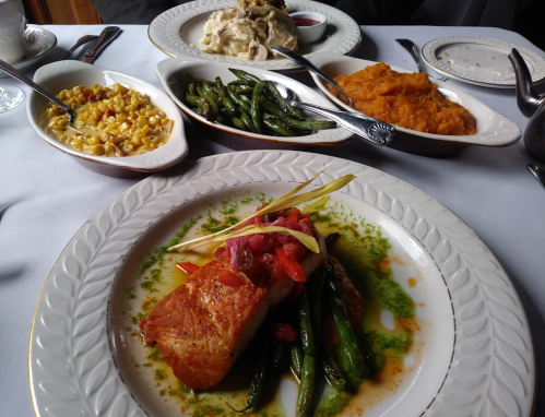 A plate of salmon with vegetables, surrounded by side dishes of macaroni, green beans, sweet potatoes, and pasta.