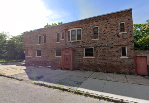 A two-story brick building with a red door, surrounded by a concrete sidewalk and overgrown vegetation.