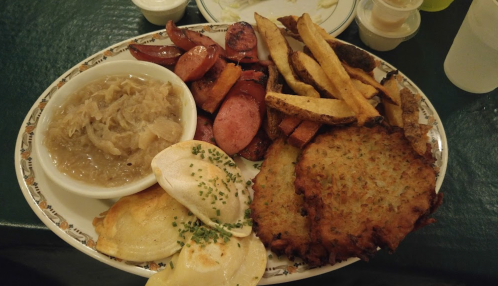 A plate of food featuring sausages, fries, sauerkraut, pierogi, and potato pancakes, garnished with chives.