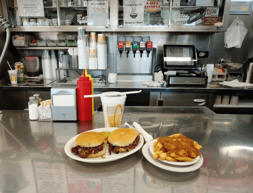 A diner counter with two burgers, fries, and a drink on a plate, with condiments and a soda fountain in the background.