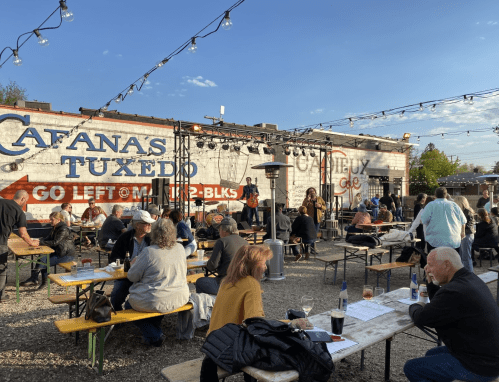 Outdoor venue with people dining and enjoying live music under string lights, with a mural in the background.