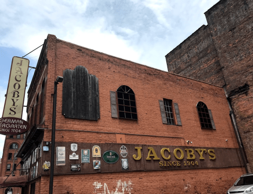 A brick building with the sign "Jacoby's Since 1904" and various plaques on the wall, under a cloudy sky.